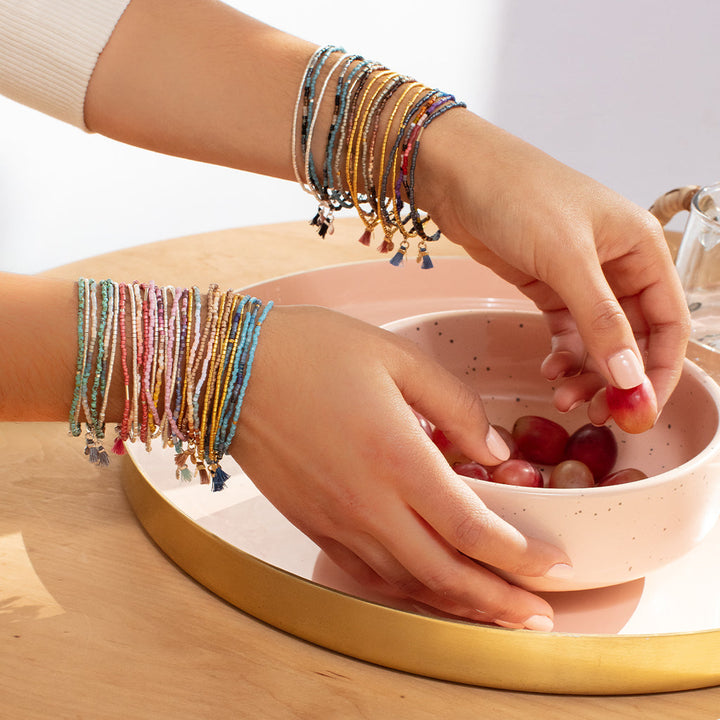 Two hands adorned with the Turquoise Multi Silver Miyuki Bracelet Trio by Scout Jewelry, made from vibrant Miyuki Delica beads, are reaching into a pink bowl filled with cherries on a wooden table. The table is partially covered by a golden tray. The scene is well-lit, giving a bright and cheerful atmosphere.