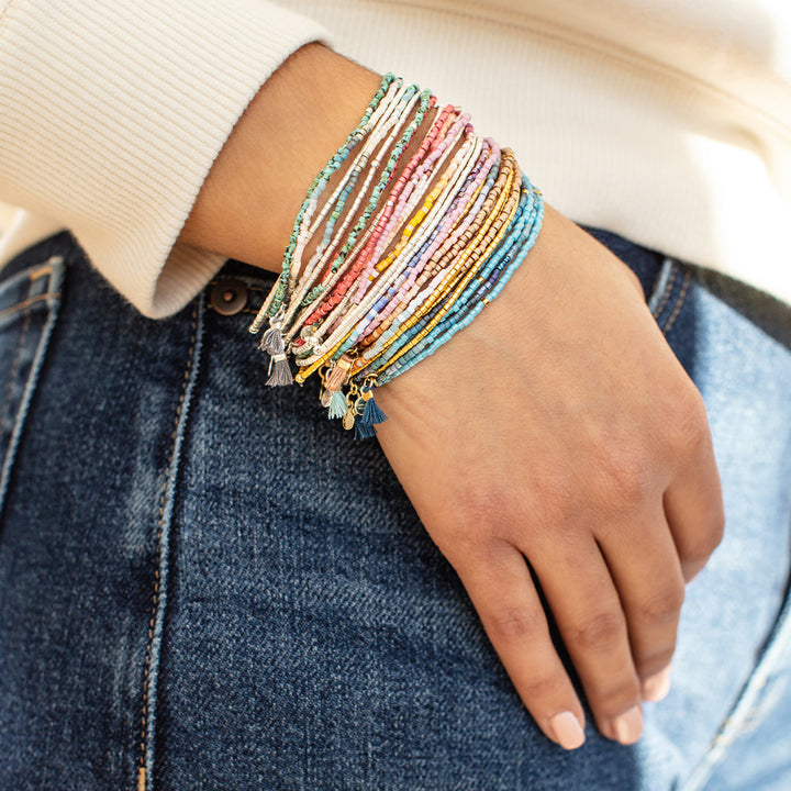 A person wearing a cream-colored sweater and denim jeans is shown with their hand resting on their hip, adorned with the Blush Multi Silver Miyuki Bracelet Trio by Scout Jewelry. The bracelets are crafted from vibrant Miyuki Delica beads, featuring tassel accents in an array of colors gracefully draped around their wrist.