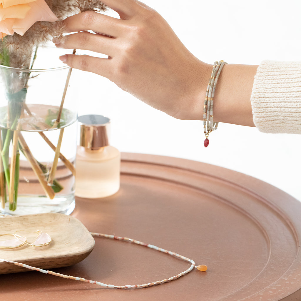 A hand reaches towards a flower arrangement in a glass vase, adorned with the Peace Aqua Terra Silver Teardrop Wrap from Scout Jewelry, featuring semi-precious gemstones and a red charm. The setup includes a perfume bottle, rose-tinted glasses, and a tiered necklace on a round tray, all on a softly lit surface.