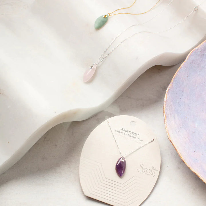 A delicate display featuring three gemstone necklaces showcases the beauty of Scout Jewelry. Two pieces rest elegantly on a marble surface: one is the Amazonite Gold Organic Stone Necklace, featuring an amazonite pendant on a 14k gold chain, while the other necklace displays a lovely pink stone. A third necklace, with a stunning purple amethyst pendant and sterling silver chain, is partially packaged on a card that reads "Scout." A ceramic dish is also nearby.