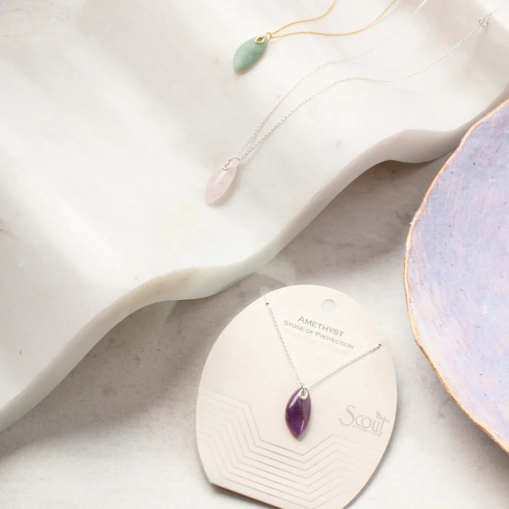 A sophisticated jewelry display showcases three necklaces on a white marble surface. The chains, elegantly crafted from 14k gold, exhibit teardrop-shaped pendants in green, pink, and purple. Notably, the necklace with the purple amethyst pendant is attached to a card labeled "Amethyst Silver Organic Stone Necklace" from Scout Jewelry. A decorative dish is partially visible in the scene.