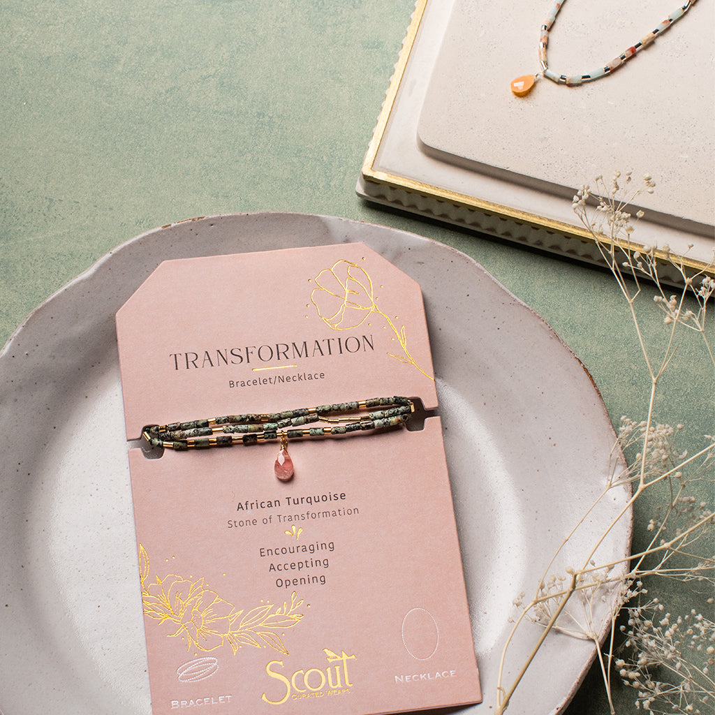 A bracelet card labeled “Peace Aqua Terra Silver Teardrop Wrap” from Scout Jewelry sits on a green surface, featuring a piece adorned with African Turquoise and a pink gem. Nearby, a similar necklace rests on a cream-colored dish. Semi-precious gemstones glisten amidst scattered dried flowers.