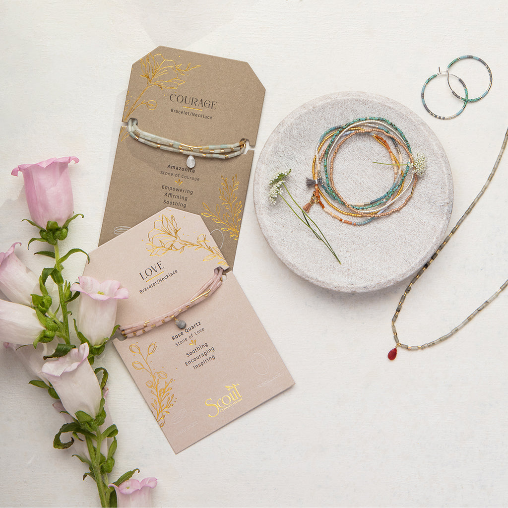 A flat lay of Scout Jewelry features the Love Rose Quartz Gold Teardrop Wrap bracelet on a "Love" card and another on a "Courage" card. A white dish holds semi-precious stone bead bracelets, including labradorite. Silver hoop earrings and pink flowers embellish the left side.