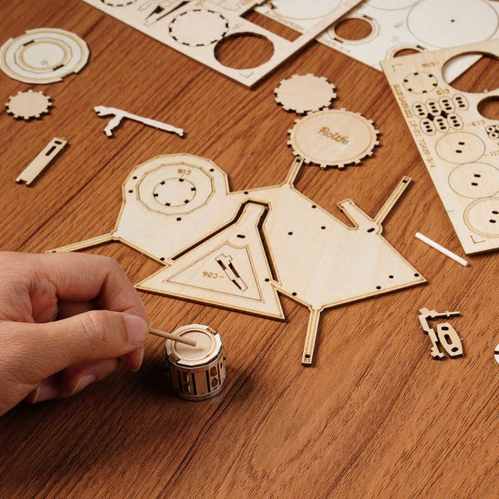 A person is meticulously assembling the Laser Cut Wooden Puzzle: Drum Kit by Hands Craft on a wooden surface. The kit features various pre-cut pieces, including shapes like circles and triangles, along with intricate components. The person's hand is holding one small piece while using a toothpick for precise assembly.