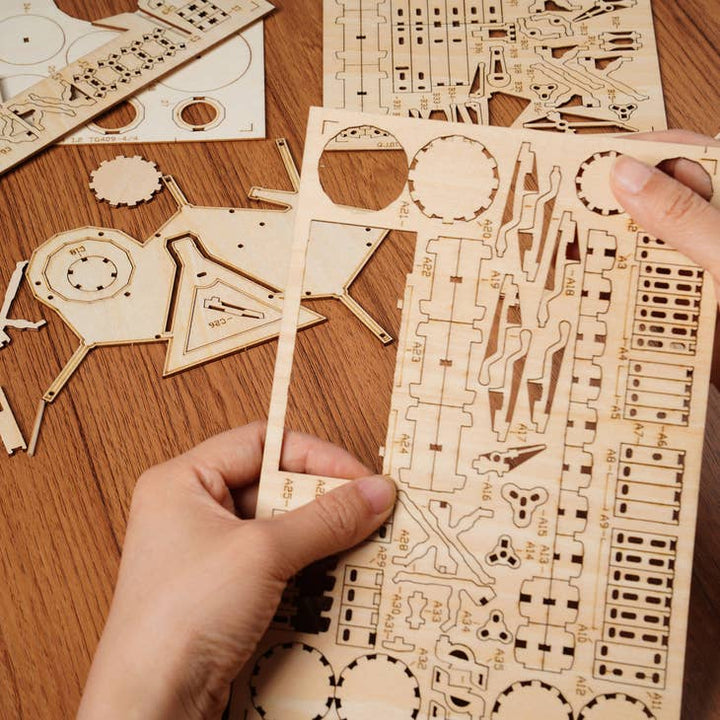 Hands meticulously assembling a Hands Craft Laser Cut Wooden Puzzle: Drum Kit on a wooden surface. Various laser-cut wooden pieces, including gears and structural components, are spread out like a 3D puzzle, ready for assembly into what promises to be a detailed drum kit—a perfect music gift for enthusiasts.