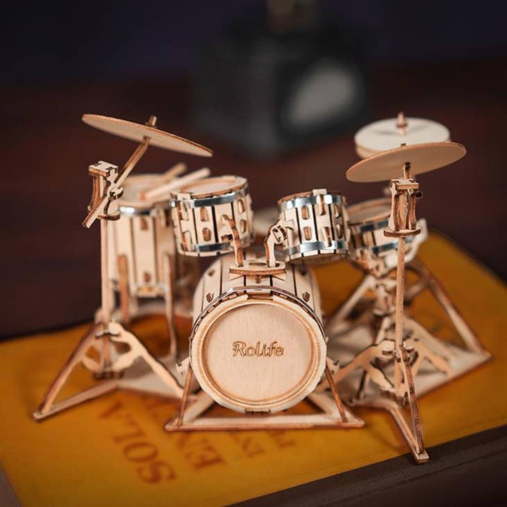 The Laser Cut Wooden Puzzle: Drum Kit by Hands Craft is an intricately detailed miniature model featuring components such as a bass drum, snare drum, tom-toms, hi-hat, and cymbals. This stunning home decoration rests gracefully on top of a book with a partially visible yellow and brown cover.