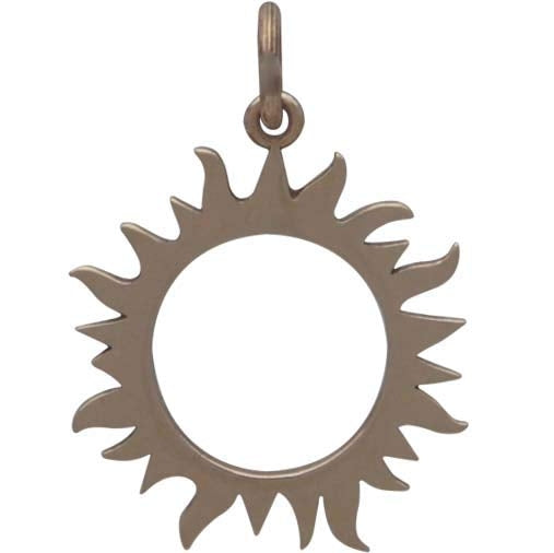 The Bronze Eclipse Sun Charm by Nina Designs is a metallic pendant resembling a solar eclipse, featuring a central cutout encircled by wavy-edged sun rays and suspended from a small loop at the top.