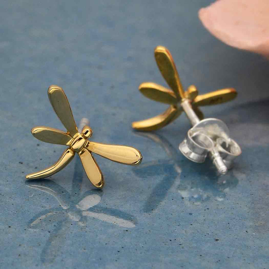 A pair of bronze dragonfly post earrings by Nina Designs is showcased on a glossy blue surface. One earring stands upright, while the other rests on its side, revealing the back and clasp. A peach-colored object is partially visible in the corner.