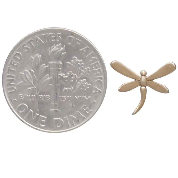 A petite golden dragonfly pin is elegantly positioned next to a U.S. dime, emphasizing its delicate size. This exquisite piece pairs beautifully with the Bronze Dragonfly Post Earrings by Nina Designs, creating a stunning ensemble.