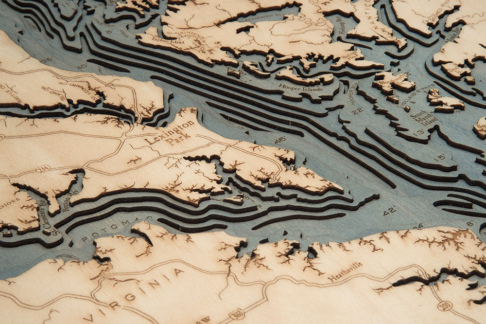A close-up of the WoodChart Chesapeake Bay 24.5" x 31" map in a white frame highlights laser-cut wood layers depicting coastal inlets, islands, and labeled locations, with contours representing water depth and land elevation.