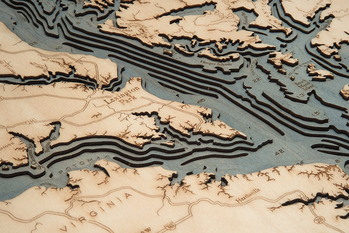 A close-up of the WoodChart Chesapeake Bay 24.5" x 31" map in a white frame highlights laser-cut wood layers depicting coastal inlets, islands, and labeled locations, with contours representing water depth and land elevation.