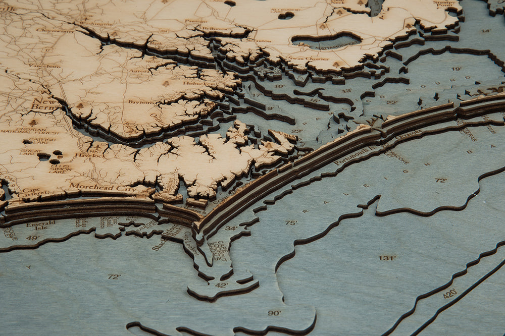 A close-up of the WoodChart North Carolina Coast 24.5" x 31" White Frame, a layered wooden topographic map featuring engraved details and contour lines of the coastline and waterways near Morehead City, North Carolina.