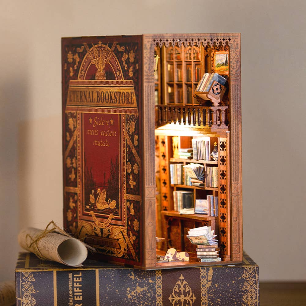 The Hands Craft DIY Book Nook Kit: Eternal Bookstore with Dust Cover opens to reveal a miniature, illuminated bookstore scene—shelves, books, intricate details—making it a unique literary gift.