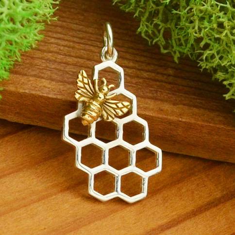 A Nina Designs Sterling Silver Honeycomb Charm with a Bronze Bee is showcased on wood, adorned by green foliage in the background.