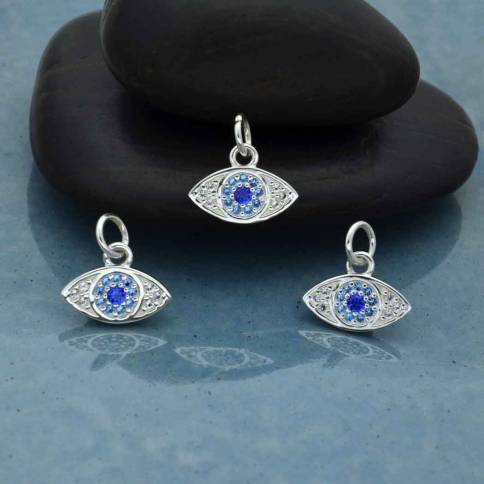 Three Nina Designs Sterling Silver Pave Set Nano Gem Eye charms rest on a smooth blue surface, two lying flat and one leaning against stacked black stones, highlighting the allure of this striking evil eye design.