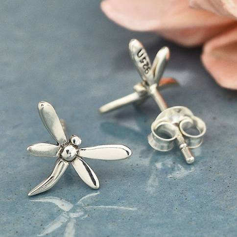 Close-up of Nina Designs' Silver Dragonfly Post Earrings on a blue-gray surface, with a soft focus pink flower in the background. The polished design reflects their spirit animal symbolism, and the "925" marking confirms their quality.