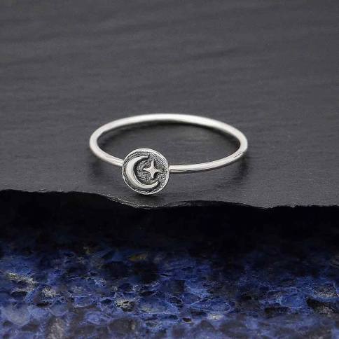 The s5 Raised Moon and Star Stacking ring by Nina Designs is a delicate sterling silver piece featuring a round moon and star motif, ideal for stacking. It is elegantly displayed on a dark stone surface with a textured blue background.