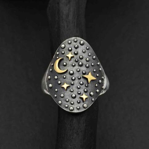 The s6 Night Sky Ring with Moon and Stars by Nina Designs, elegantly showcased on a black background, features a dark oval face adorned with a bronze moon and stars. Silver dots enhance its design, capturing the essence of a starry night sky.