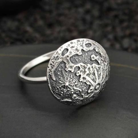 Nina Designs presents the Full Moon Silver Size 7 Ring, crafted in sterling silver and featuring a round, textured top adorned with intricate abstract designs. The smooth band elegantly contrasts with the detailed top, reminiscent of a full moon. This sophisticated piece rests beautifully on a dark, textured surface.