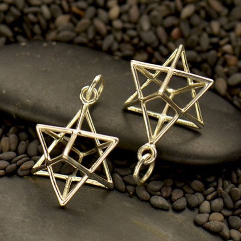 Two Sterling Silver Wire Merkaba Pendants by Nina Designs rest on smooth black stones, surrounded by small black pebbles.