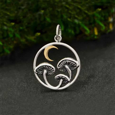 The Mushroom Charm with Bronze Moon by Nina Designs showcases a circular pendant adorned with three mushrooms featuring gracefully curved stems. A bronze crescent moon elegantly sits in the upper left section of the circle, while a backdrop of lush forest greenery enhances its captivating charm. This enchanting mushroom charm is suspended by a small loop at the top.