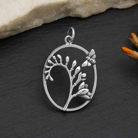 Nina Designs presents the Bee and Freesia Flower Pendant, a sterling silver piece with an oval frame embellished by a delicate leaf branch and a small bee perched on the side. The background displays textured stone and subtle decorative elements, creating an enchanting pendant that celebrates nature's beauty.