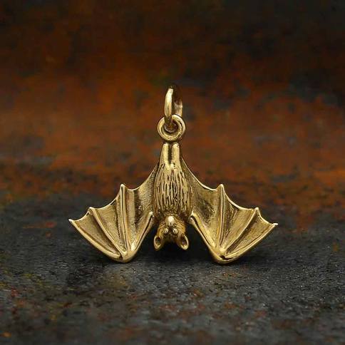 The Bronze Realistic Bat Pendant by Nina Designs features a meticulously detailed bat with intricate wings and body, suspended upside down to accentuate its realistic appearance. Set against a textured dark backdrop, the pendant's metallic sheen stands out, embodying the symbolic essence of bats.