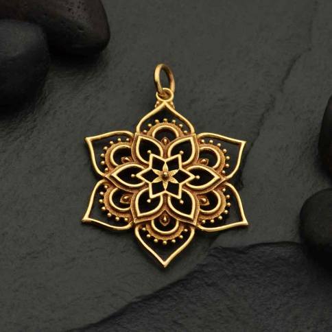 The Bronze Openwork Lotus Mandala Pendant by Nina Designs rests on a dark textured stone surface, encircled by smooth black pebbles.