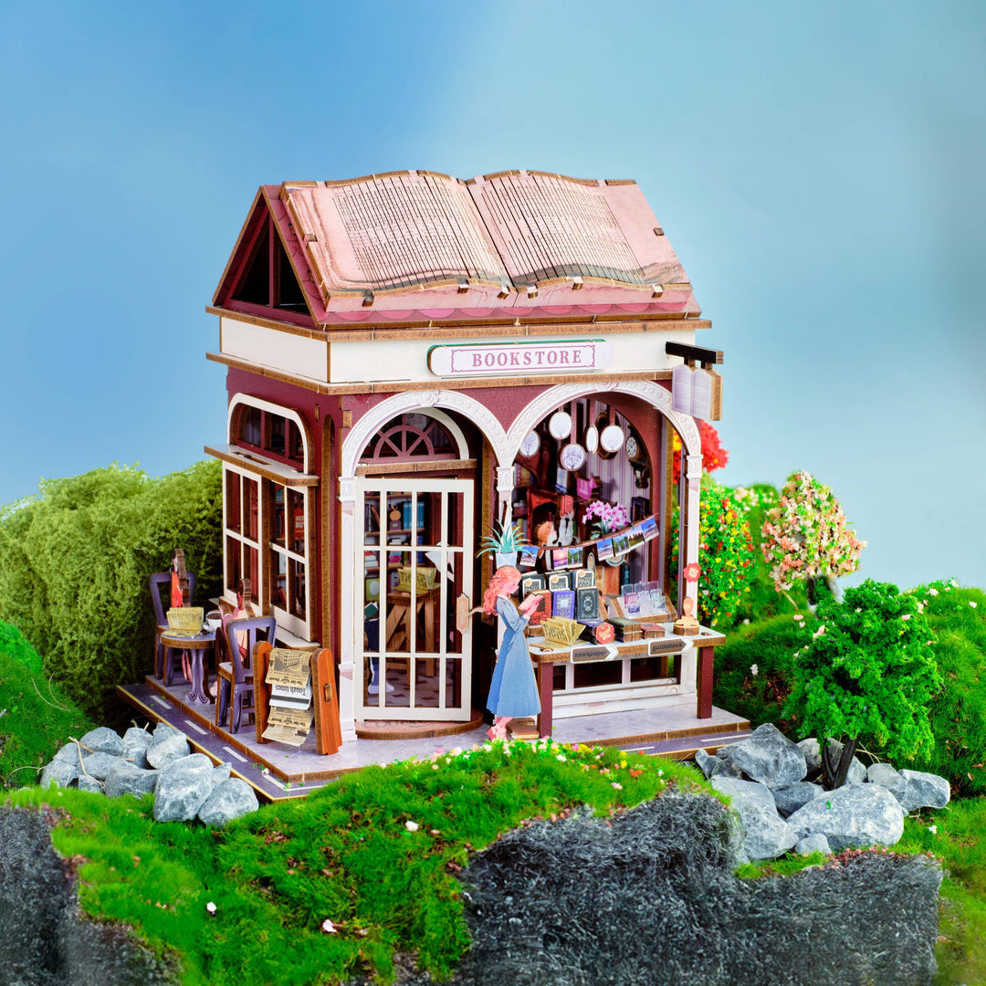 Create a charming book nook with the Hands Craft DIY Miniature House Kit: Bookstore, featuring a cozy cottagecore design with a large book-shaped roof, outdoor seating, colorful decor, greenery, rocks, and a small tree.