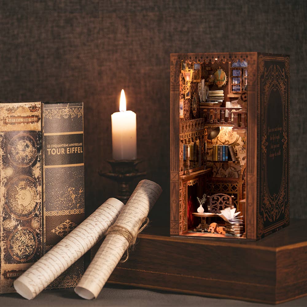 A candle, antique books, and scrolls sit beside the Hands Craft DIY Book Nook Kit: Eternal Bookstore with Dust Cover, featuring intricate miniature shelves, a desk, and warm lighting—perfect for creating a cozy library scene or unique literary gift.