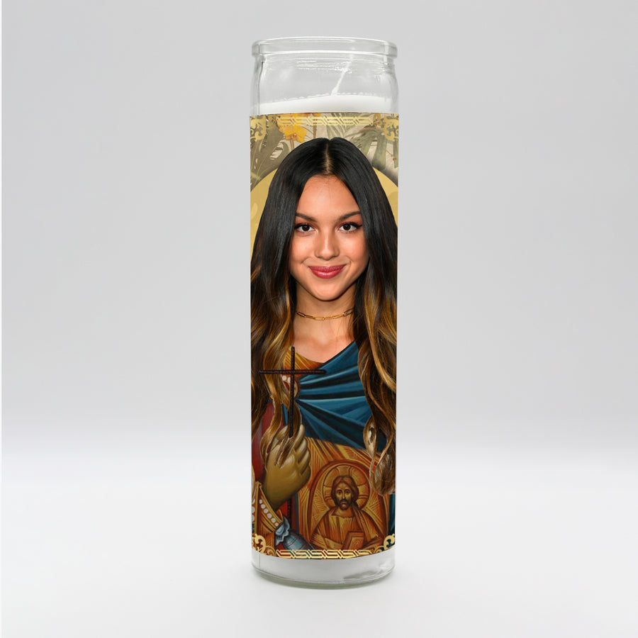 The Olivia Rodrigo Candle by BOBBYK BOUTIQUE is a tall, unscented candle showcasing an artistic depiction of a woman with long dark hair in religious iconography style, featuring a halo and ornate decorations, including smaller religious figures at the base. Perfect for any setting.