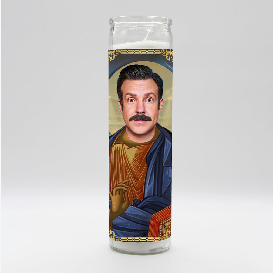 The Ted Lasso Candle by BOBBYK BOUTIQUE is a tall devotional candle featuring a stylized saint-like portrait of a mustachioed man resembling Ted Lasso. Dressed in a blue robe, he holds a book and an object resembling a heart, adorned with ornate gold accents. The background includes a halo effect, enhancing the holy iconography.