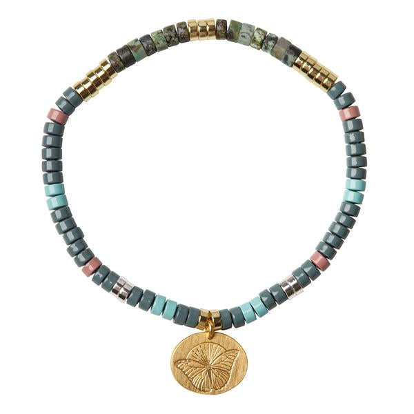 The "Intention Charm African Turq/Gold" bracelet by Scout Jewelry features a beaded stretch cord with small, square beads in shades of teal, gray, green, and gold. It also includes a few pink beads and a single round gold charm engraved with a butterfly design. This positive intention charm adds a meaningful touch to the semi-precious stones.