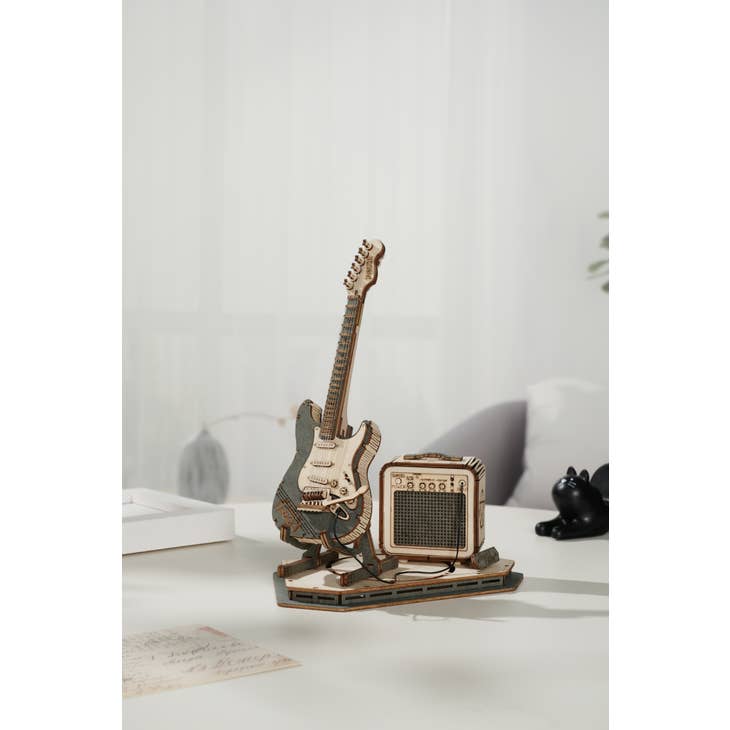 A detailed, laser-cut wooden puzzle of an electric guitar from Hands Craft's Musical Instrument Series and a small amplifier sit on a desk. The background is softly blurred, showcasing a minimalistic and modern interior. A black cat figurine and a piece of paper are partially visible in the background.