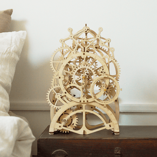 The DIY Pendulum Clock from Hands Craft, featuring intricate gears and wheels, sits on a dark wooden surface beside a white pillow. This wooden mechanical design is a 3D puzzle with interlocking parts, showcasing exquisite craftsmanship and complexity.