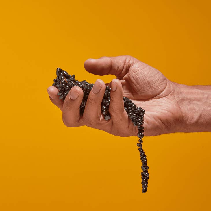 A hand holds SPEKS Crags Magnetic Putty Stones—shiny black magnets—against a solid bright orange background, ideal for stress relief.