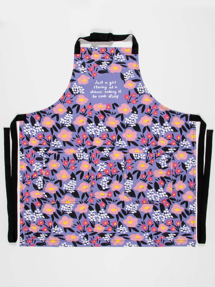 Introducing the "Just a girl Apron" by Blue Q: This apron is crafted from vibrant cotton twill, featuring a floral pattern with yellow, pink, and purple flowers nestled among black leaves. It includes an easy-adjust neck strap and extra-long waist ties for added comfort. The apron is adorned with the sleek white lettering that reads, "Just a girl standing in a kitchen asking to cook dinner.