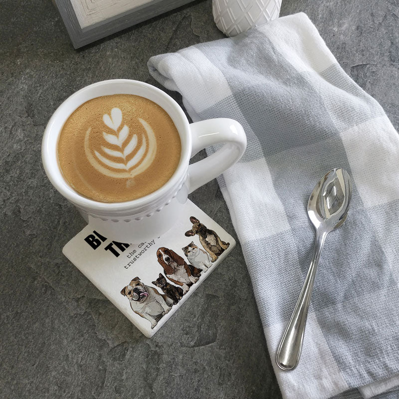 A cup of coffee adorned with latte art rests on a "Beware of Dog Coaster" by Tipsy Coaster, known for its absorbent design featuring dog images. Beside it, a silver spoon and a checkered cloth lie on the gray countertop.