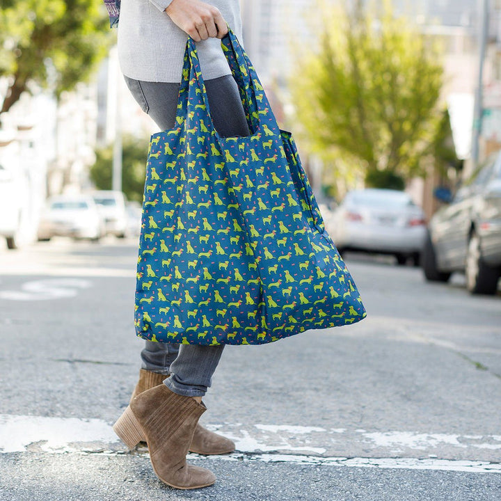 A person walks across the street with a Dog Pack Reusable Bag by zzzRockFlowerPaper, which features an environmentally friendly design adorned with a yellow animal pattern. They sport gray jeans and beige ankle boots, while parked cars and blurred trees create the backdrop.