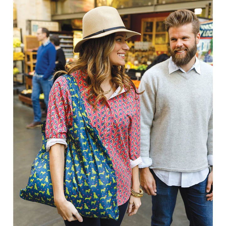 A woman sporting a hat and a patterned shirt beams as she walks alongside a man in a gray sweater. She holds an environmentally friendly, vibrant Dog Pack Reusable Bag by zzzRockFlowerPaper. They're in a market environment with produce and other shoppers visible in the background.