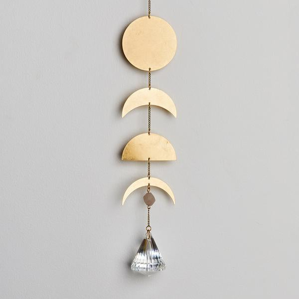 The Moon Phase Moonstone Suncatcher by Scout Jewelry features a decorative hanging with geometric shapes in gold, including a circle, two semicircles, and a small crescent. A faceted crystal hangs below, casting rainbows when it catches the light against a neutral wall.