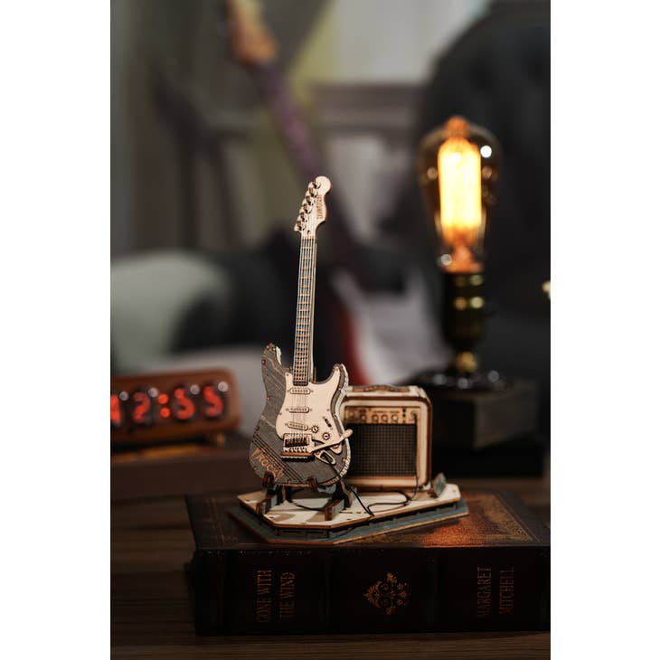 The Laser Cut Wooden Puzzle: Electric Guitar from Hands Craft's collection is showcased on a book with vintage styling. Behind it, an antique-style light bulb glows warmly, and a digital clock indicates the time as 12:55. The background features a blurred room setting adorned with various objects.