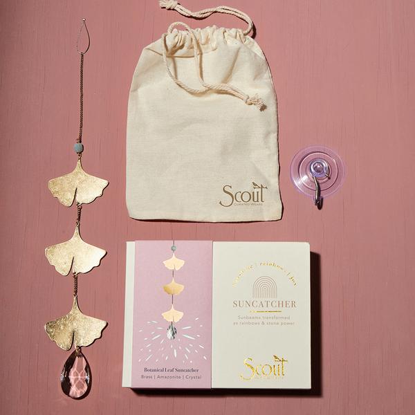 Set against a pink background, the Moon Phase Moonstone Suncatcher by Scout Jewelry features gold leaves and pink semi-precious gemstones. It comes with a beige pouch labeled "Scout" for safekeeping and is packaged in a box. Additionally, a suction cup is included to create dancing rainbows across your space.