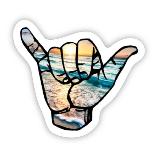 The Hang Loose Photo Sticker by Big Moods features a stylized hand making a "shaka" sign (thumb and pinky extended, other fingers curled), filled with an ocean scene during sunset. The background showcases a vibrant, colorful sky and waves crashing onto the shore. This high-quality sticker has a white border and embodies captivating coastal beauty.