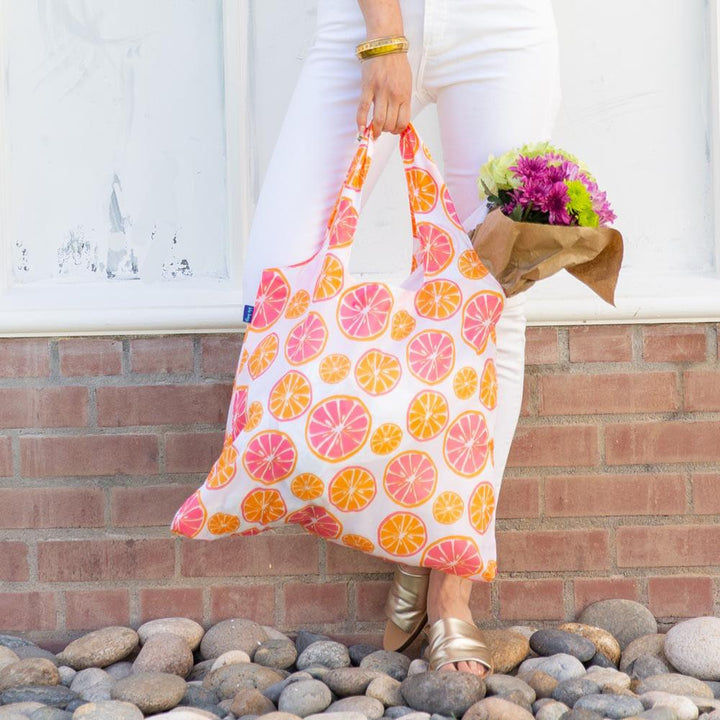 A person in white pants holds a zzzRockFlowerPaper Citrus Reusable Bag adorned with vibrant citrus fruit patterns. They are wearing gold sandals and a yellow bracelet, while carrying a bouquet of flowers wrapped in brown paper. The backdrop consists of a charming brick and white wall.