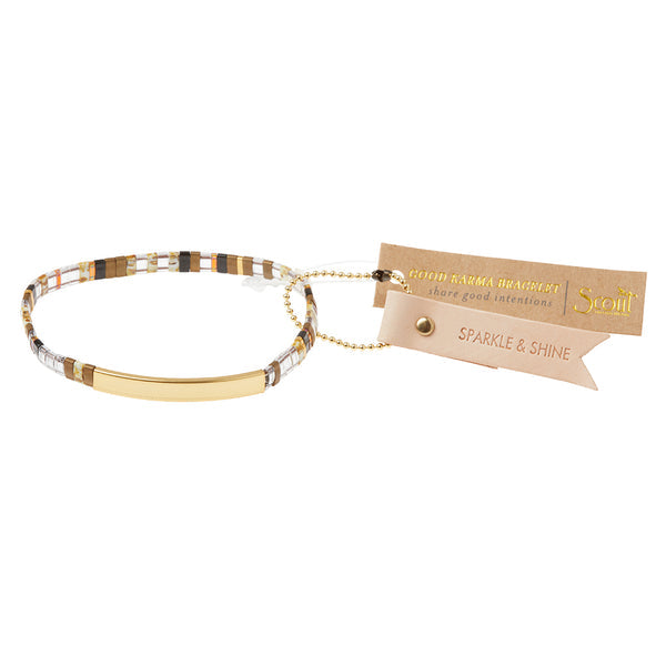 The Miyuki Sparkle and Shine Topaz Gold bracelet by Scout Jewelry features a 14K gold-dipped bar and a tag that reads "SPARKLE & SHINE." It is crafted with Japanese glass Miyuki beads in elegant shades of white, gold, and brown, and includes a tan tag that says "GOOD KARMA BRACELET share good intentions.