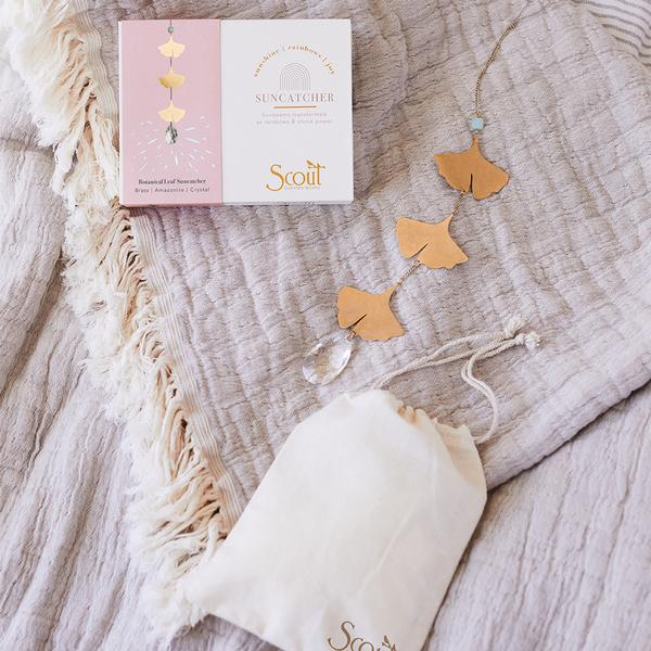 A delicate Leaf Amazonite Suncatcher with a crystal casts rainbows across a textured beige blanket, next to an open package labeled "Scout Jewelry." A cream drawstring bag adorned with semi-precious gemstones lies beside them.