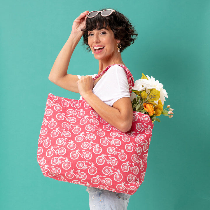 A smiling person with short hair holds the Beach Cruiser Jute Tote, a large red bag from zzzRockFlowerPaper, adorned with white bicycle patterns. The biodegradable tote is filled with white and yellow flowers. They stand against a turquoise background, wearing a white shirt and sunglasses on their head, embracing sustainable products.