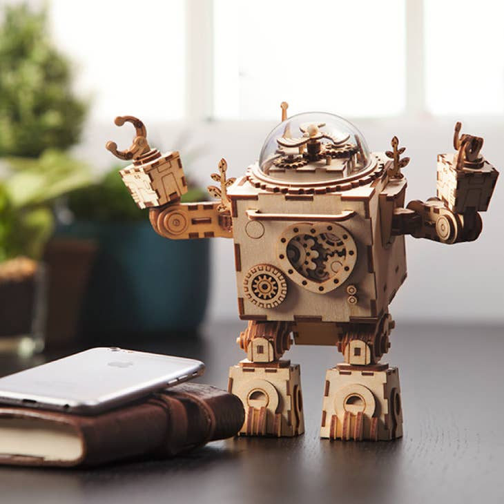 A DIY 3D Steam Punk Music Box featuring Orpheus by Hands Craft, adorned with gears and intricate details, sits on a table beside a closed book and a smartphone. The music box showcases a domed head and articulated arms, set against a blurred background with plants and a window to evoke a subtle steampunk aesthetic.