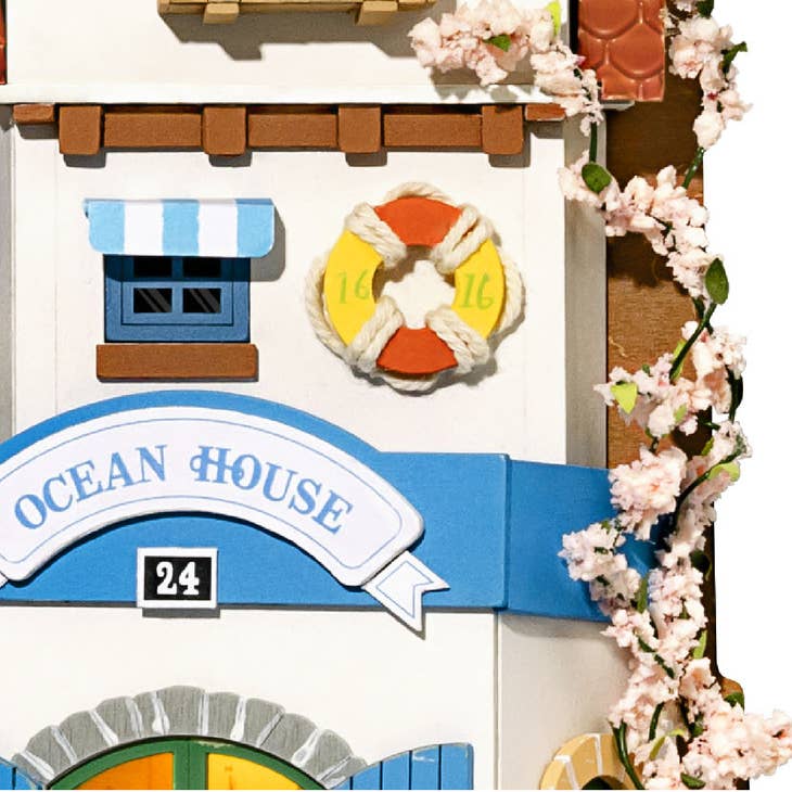 The DIY Miniature Wall Hanging Kit Island Dream Villa by Hands Craft makes for a unique gift idea for crafters and collectors. This decorative piece resembles a charming house and features a lifebuoy, an "Ocean House" sign, blue windows, and a striped awning. Cherry blossom decorations add a natural touch, with the number "24" displayed below the sign.